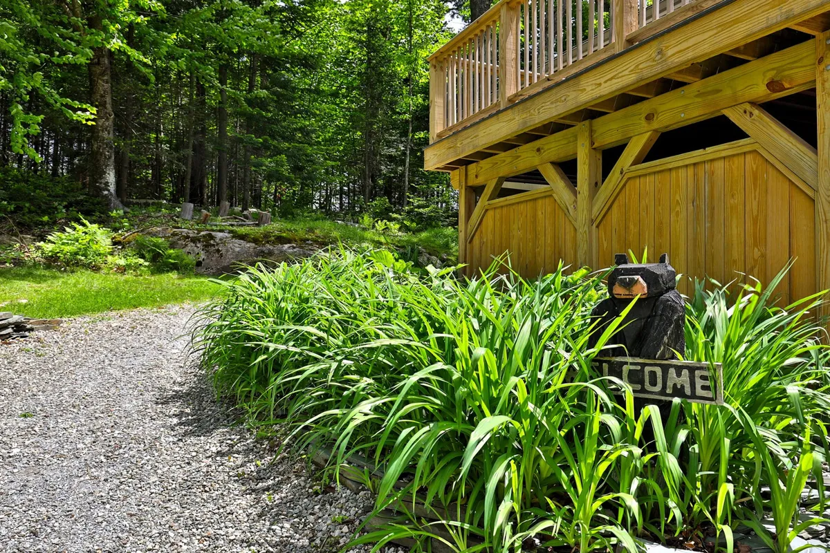 The photo captures the welcoming entrance of the property, featuring a wooden deck adorned with a bear welcome sign and surrounded by lush greenery. This inviting scene sets the mood for tranquility and connection with nature.