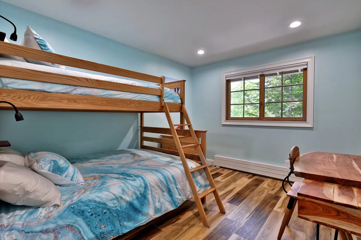 This cheerful bunk room includes a twin-over-full bunk bed with colorful bedding, a wooden desk and chair, and bright natural light streaming through a window. The space is playful yet cozy, ideal for kids or friends.