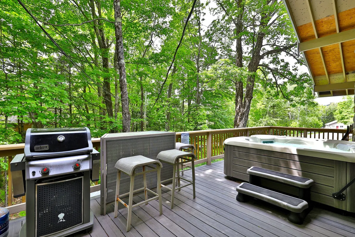 A relaxed outdoor setting featuring a hot tub, grill, bar stools, and the surrounding trees. The scene feels inviting and offers a glimpse of après ski relaxation under the open sky.