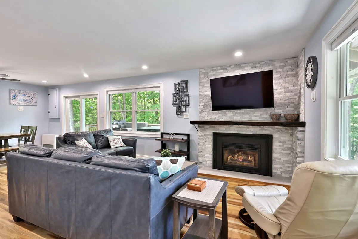This image features a comfortable leather sofa positioned near a glowing fireplace, with large windows letting in natural light and a smart TV mounted on the wall. The arrangement is perfect for family gatherings, relaxing by the fire, or enjoying a movie night.