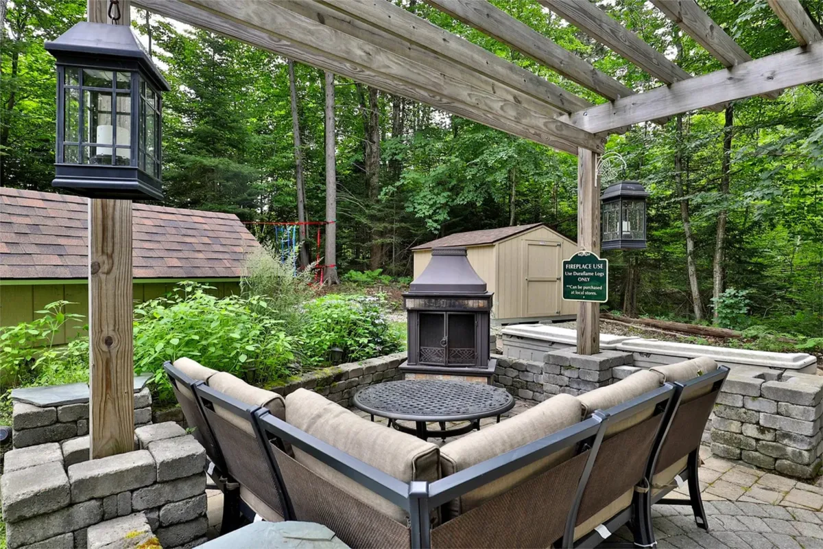 A cozy exterior area with a pergola-covered seating arrangement, outdoor fireplace, and lush garden greenery. The setup invites socializing and fireside chats under the open sky, perfect for group gatherings or evening relaxation.