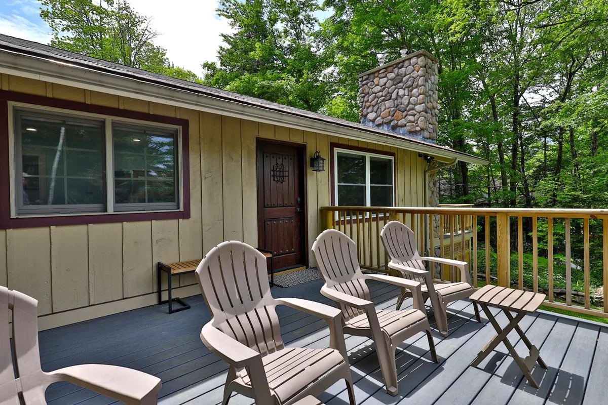 A welcoming wooden deck at the front of the house, featuring Adirondack chairs positioned for relaxation and a view of the surrounding woods. The stone chimney and front door add homey character, while the deck is spacious enough for gatherings or quiet mornings.