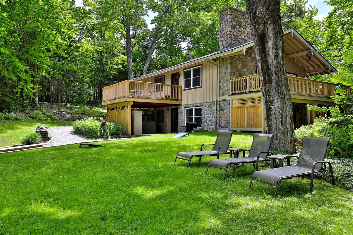 This exterior image features the property’s stone chimney, a welcoming balcony, a spacious green lawn, and a comfortable seating area. The setting is peaceful, with hints of sunset lighting and plenty of space for outdoor relaxation or gathering with friends.