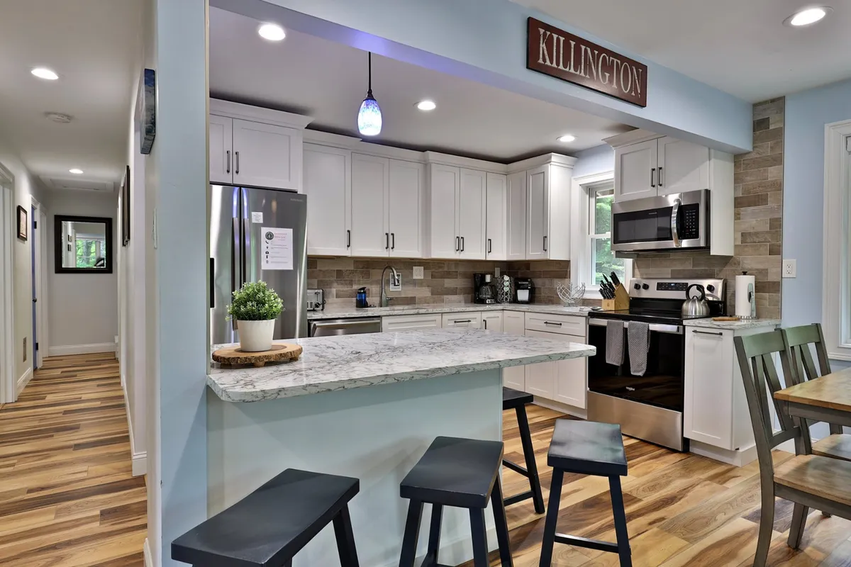 This image showcases a fully renovated kitchen with a spacious island and seating, gleaming granite countertops, stainless steel appliances, and a wooden dining table nearby. The setup is open and integrates seamlessly with the rest of the living space.