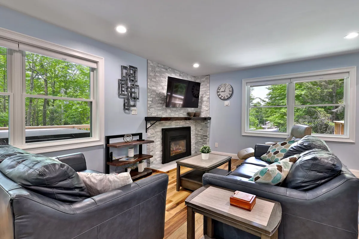 The image shows a warm, inviting living room with a crackling fireplace as the centerpiece, surrounded by a plush sofa and coffee table, with large windows framing tranquil wooded views. The space is designed for comfort and socializing, perfect for relaxing after a day on the slopes.