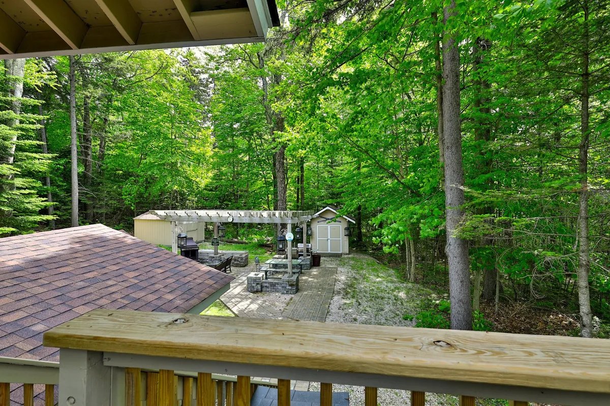 This image features a scenic view from the deck, showcasing an outdoor seating area with comfortable chairs, a pergola, and a shed surrounded by lush trees. The vantage point emphasizes the connection between the home and its tranquil wooded setting, perfect for relaxation and gatherings.