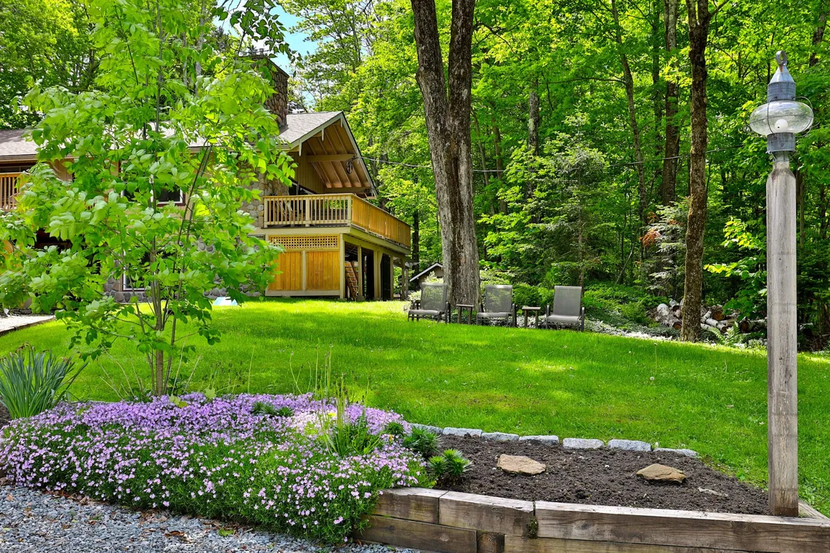 An inviting exterior shot of the house featuring a balcony adorned with flowers, lawn chairs set up on the grass, and a lamp post adding to the welcoming atmosphere. The garden setting hints at outdoor relaxation and group gatherings, with ample space for family-friendly activities.