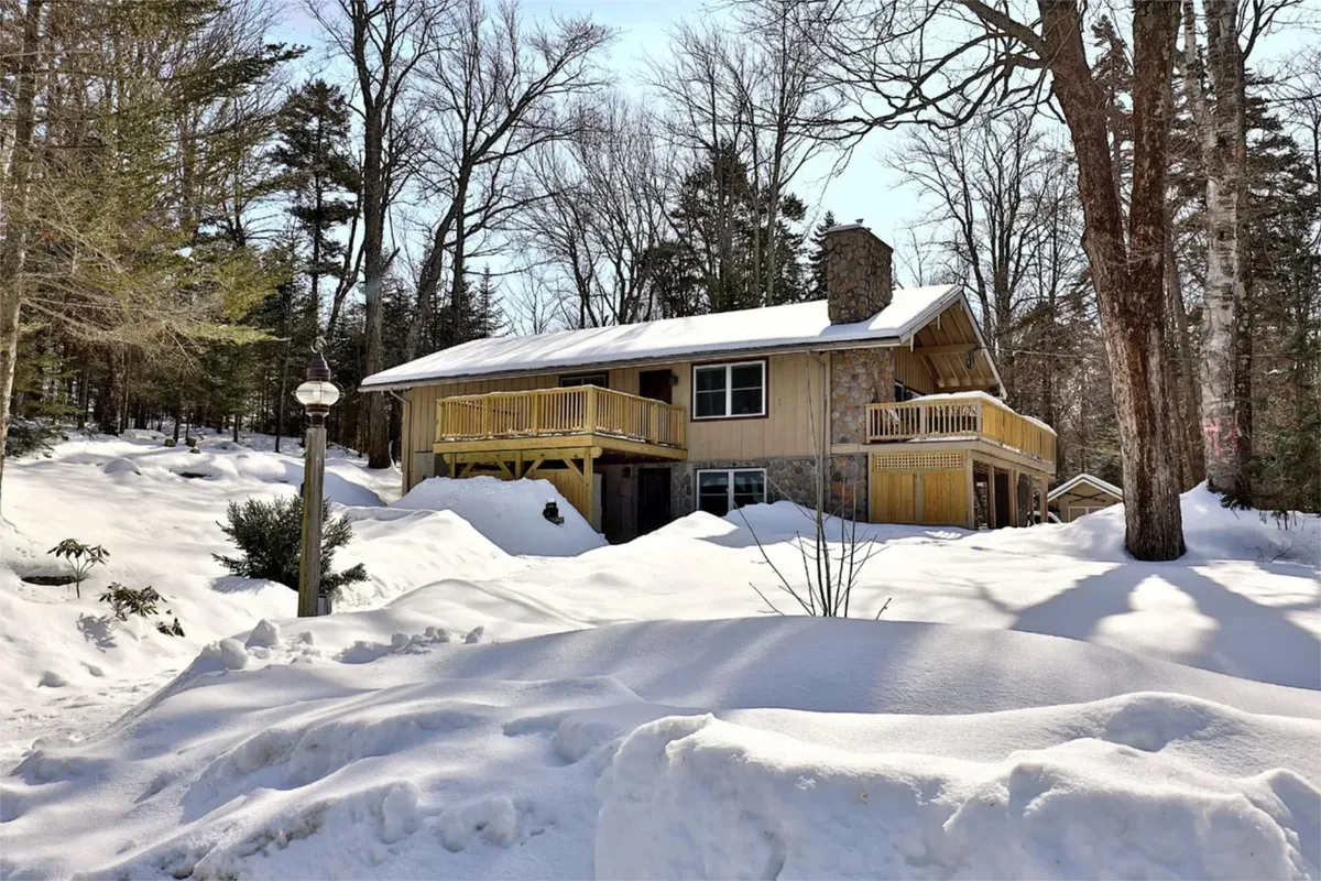 The image shows the exterior of the house set against a backdrop of snow-covered trees. The balcony and the stone chimney stand out, while fresh snow blankets the yard, evoking a classic winter getaway vibe.