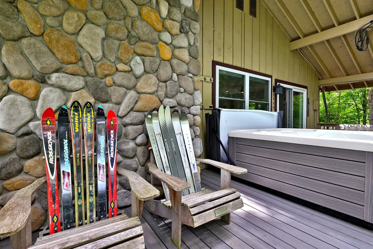 Set on an exterior deck, this image features a hot tub beside wooden chairs and skis, with a stone wall and natural surroundings. The presence of skiing gear and cozy seating highlights both post-ski relaxation and social opportunities.