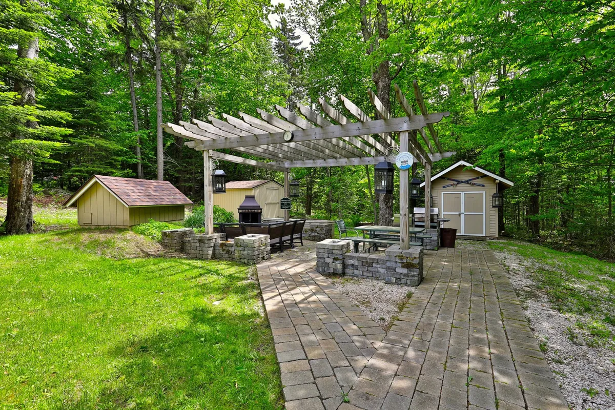 A tranquil garden scene with a pergola, an outdoor seating area, a grill, and a garden shed, surrounded by greenery. The space is set up for barbecuing, relaxing, and enjoying meals together amid nature.